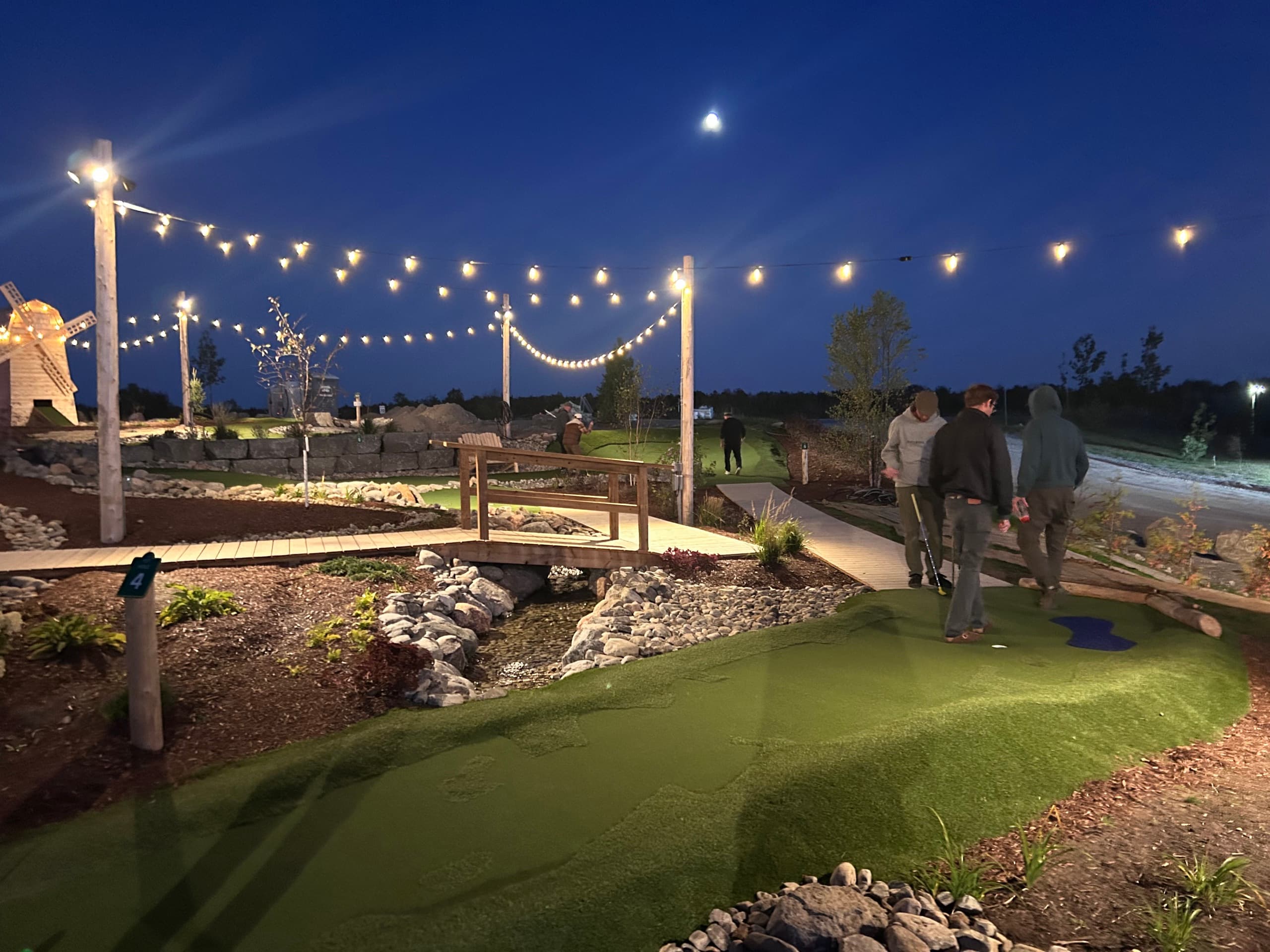 Friends playing The Little Mini Golf Co at night under string lights, with the windmill lit up